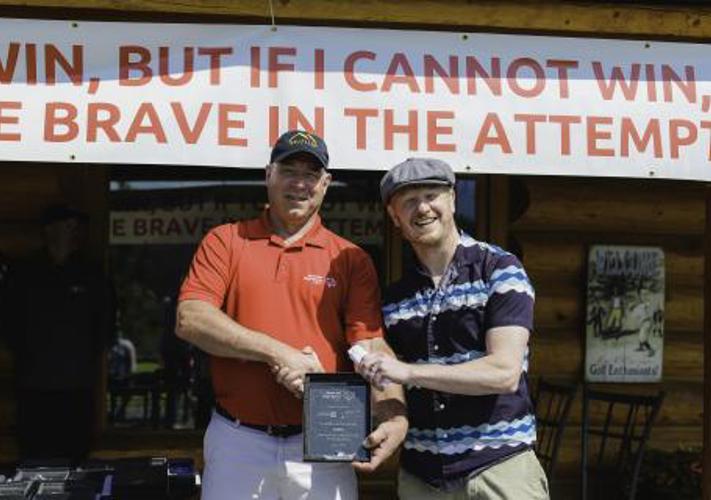 Two men are shaking hands while smiling for the camera. The man on the left is wearing a red Special Olympics Yukon shirt and is holding a plaque. The man on the right is wearing a striped shirt. Both men are standing in front of a building. Draped on the building is a sign with the Athletes Oath written on it.