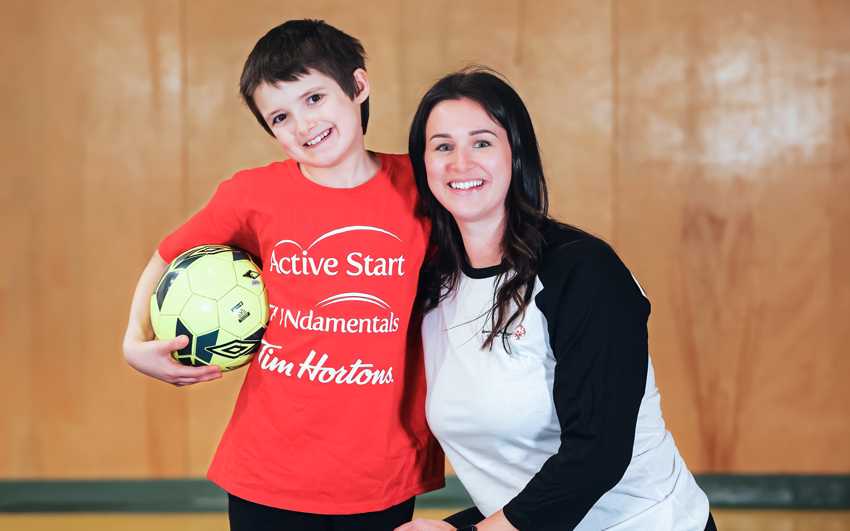 Un jeune garçon aux cheveux foncés, vêtu d'un t-shirt rouge Active Start et d'un pantalon noir, tient un ballon de football. Une femme aux longs cheveux foncés, vêtue d'un t-shirt noir et blanc et d'un pantalon noir, s'agenouille à côté de lui. L'arrière-plan est un gymnase.