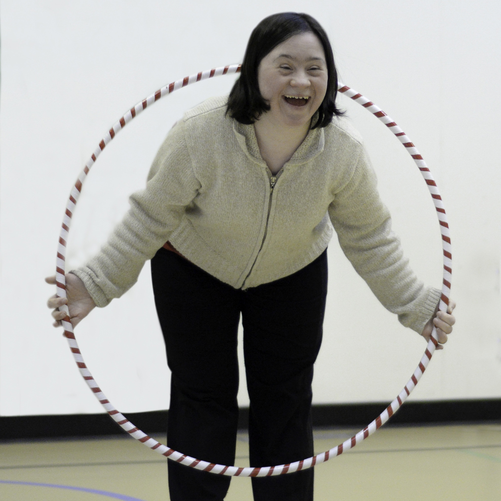 A gymnast is smiling and posing for the camera. She is wearing a white sweater and black pants, and she's holding a hula hoop. She has the upper half of her body through the hula hoop.