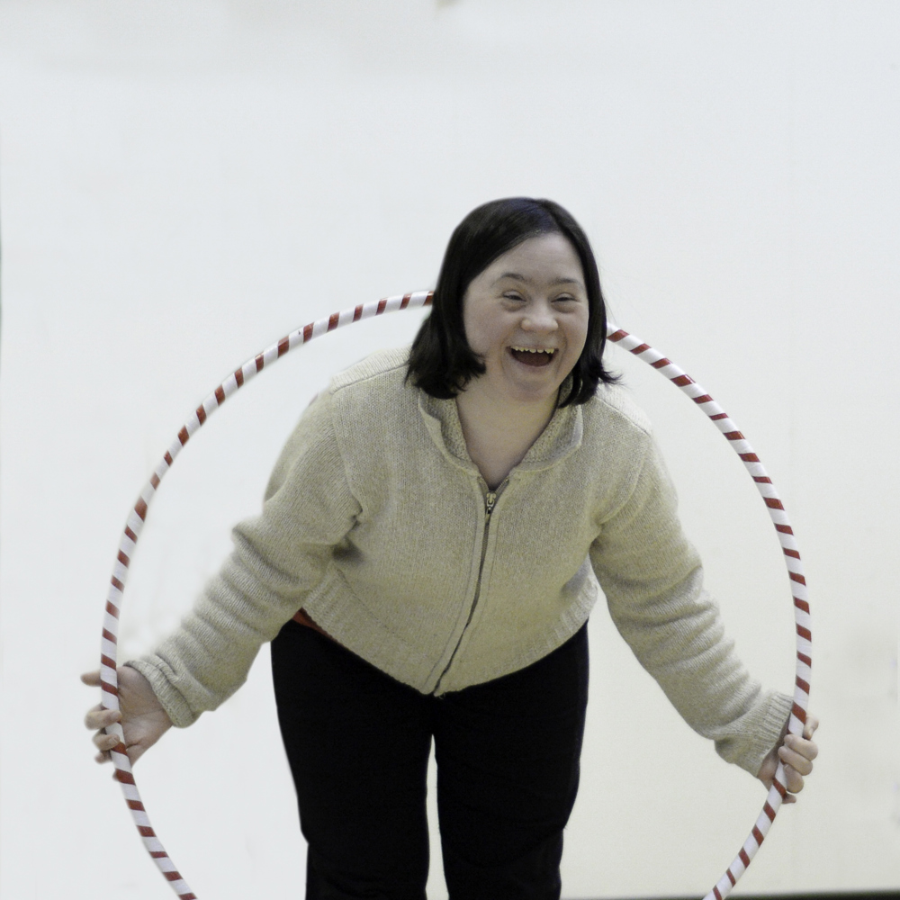 A gymnast is smiling and posing for the camera. She is wearing a white sweater and black pants, and she's holding a hula hoop. She has the upper half of her body through the hula hoop.