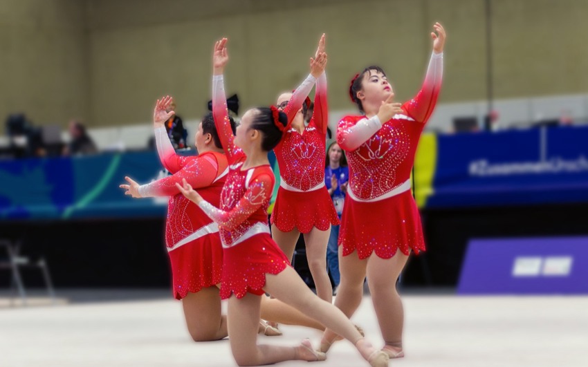 Five gymnasts wearing Special Olympics Team Canada uniforms perform a synchronized routine on the world stage, captured mid-movement with focus and energy. Representing their country with pride during an international competition.