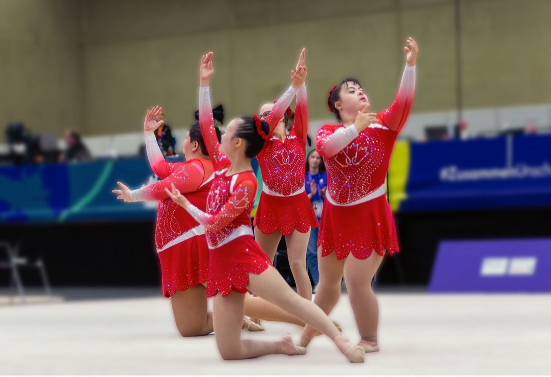 Five gymnasts wearing Special Olympics Team Canada uniforms perform a synchronized routine on the world stage, captured mid-movement with focus and energy. Representing their country with pride during an international competition.