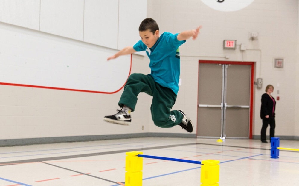 A young athlete in a teal blue t-shirt is jumping over a hurdle in a gymnasium.