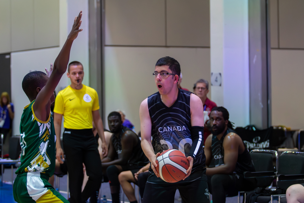 An action shot photo of a basketball game. An athlete in a "Canada" basketball jersey has possession of the basketball. An athlete on the opposing team is trying to defend their team. In the background you can see a referee with a whistle in his mouth as well as some other players on the bench.