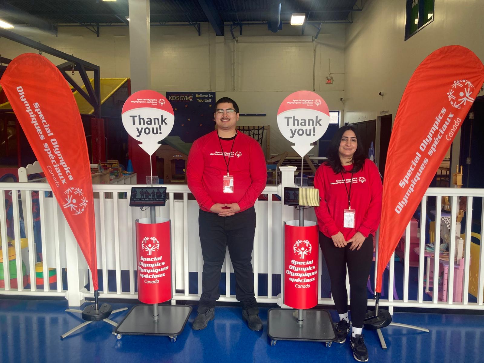 Two people in red shirts standing in front of Special Olympics signs and flags Two people in red shirts standing in front of Special Olympics signs and flags