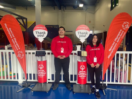 Two people in red shirts standing in front of Special Olympics signs and flags 