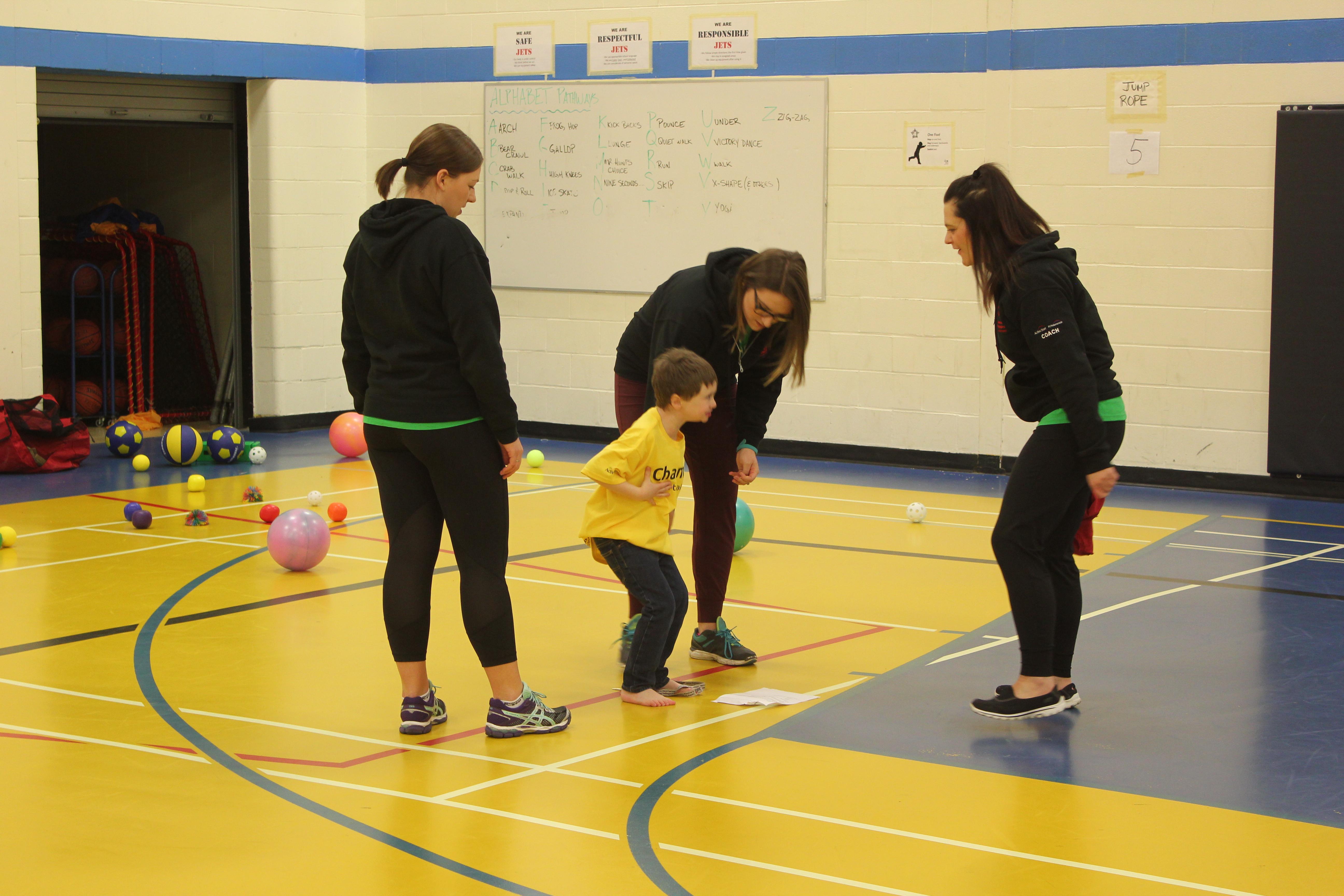Three volunteers and coaches are standing around a young athlete, assisting him with the activity. They are all standing in a gymnasium and a variety of different sized and coloured balls can be seen in the background.