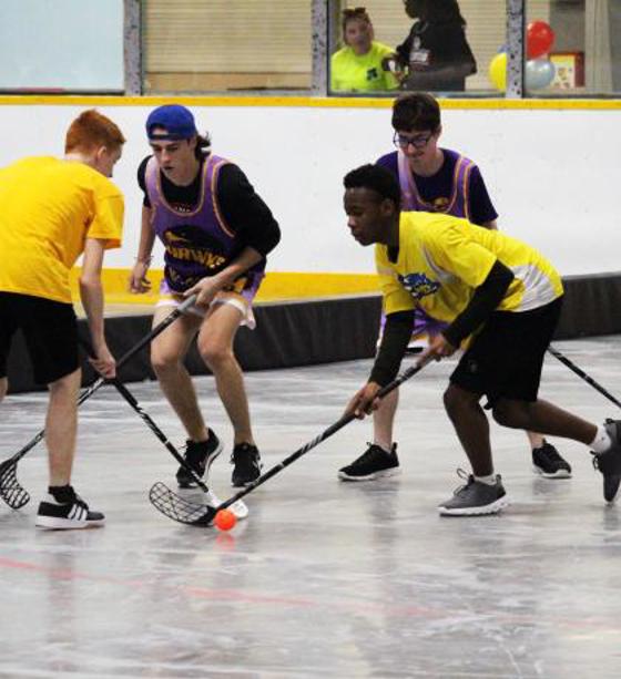Four athletes, two in yellow shirts and two in black shirts. All have on black shorts. Each is holding a hockey stick and there is an orange ball on the floor. 