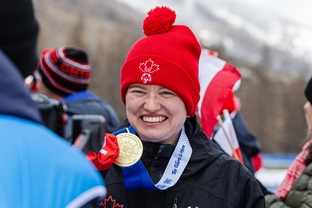 Une image en gros plan d'une athlète souriante devant une caméra qui est hors focus. Elle tient la médaille d’or suspendue autour de son cou. Elle porte également une tuque rouge et des mitaines, ainsi qu’un manteau d’hiver noir.