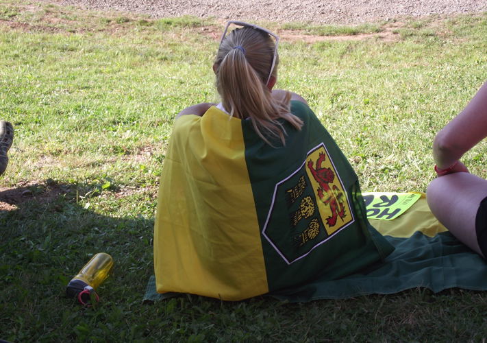 A girl is sitting on the grass with her back towards the camera. Draped around her like a cape is the Saskatchewan flag.