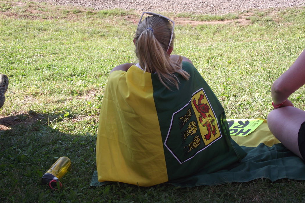 A girl is sitting on the grass with her back towards the camera. Draped around her like a cape is the Saskatchewan flag.