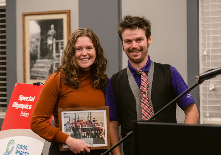 A woman in a gold long sleeved shirt and a man in a purple shirt and black vest with a red striped tie, smile and pose for the camera. They are standing next to a podium. The woman is holding a picture in a picture frame out to the camera. The picture in her frame depicts a team posing for the camera.