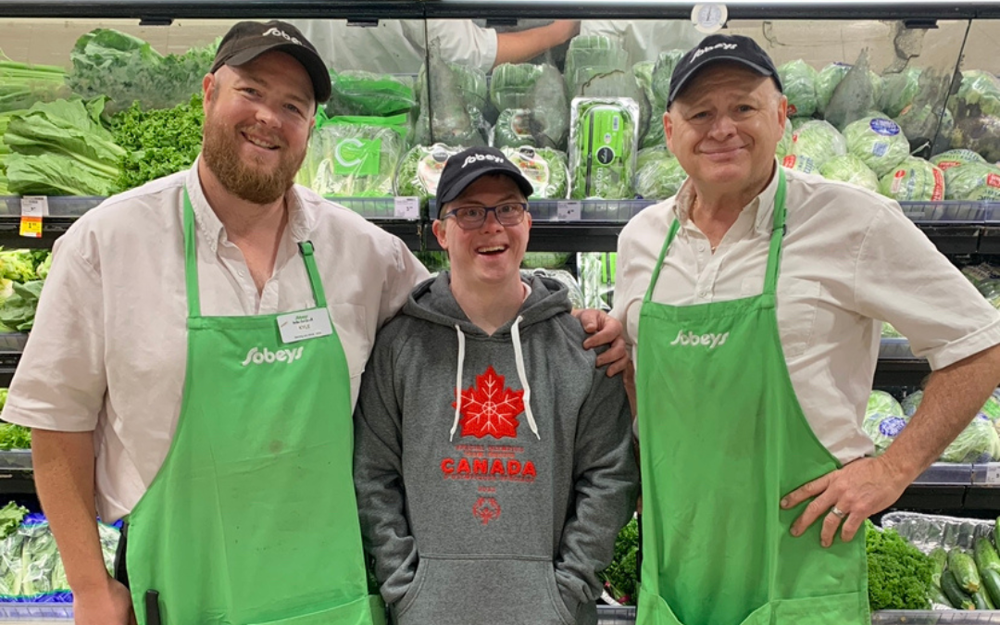 Trois hommes se tiennent côte à côte. Les deux hommes aux extrémités portent des chemises blanches et des tabliers verts avec le logo Sobeys. L'homme au milieu porte un grand sweat à capuche avec le logo du Canada. Tous sourient à la caméra. 
