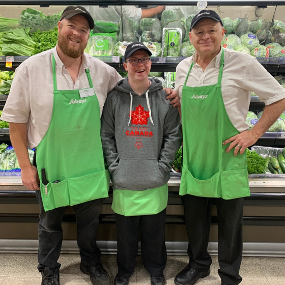 Trois hommes se tiennent côte à côte. Les deux hommes aux extrémités portent des chemises blanches et des tabliers verts avec le logo Sobeys. L'homme au milieu porte un grand sweat à capuche avec le logo du Canada. Tous sourient à la caméra. 