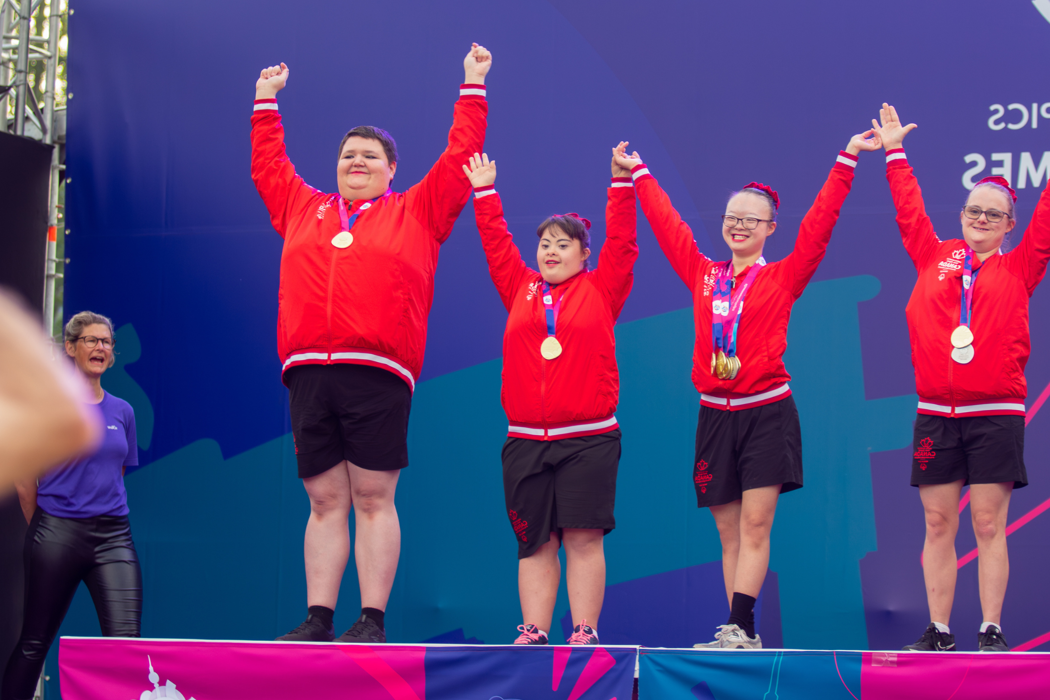Un groupe hétéroclite de personnes vêtues de rouge se tient sur un podium. Toutes ont des médailles autour du cou et les bras levés.