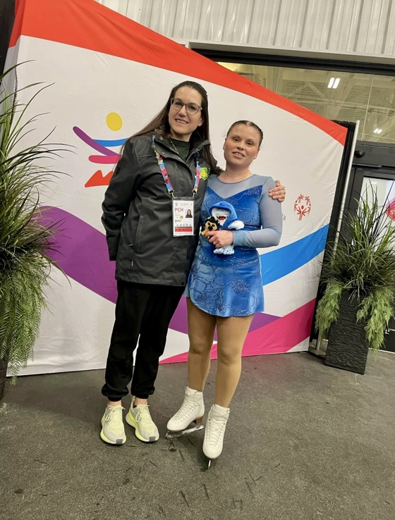 A young figure skating athlete is posing with her coach. They are both smiling for the camera and standing in front of a backdrop. The figure skater is still in her skates and blue figure skating outfit. The coach is wearing black pants and a black jacket with a colourful lanyard hanging around her neck.