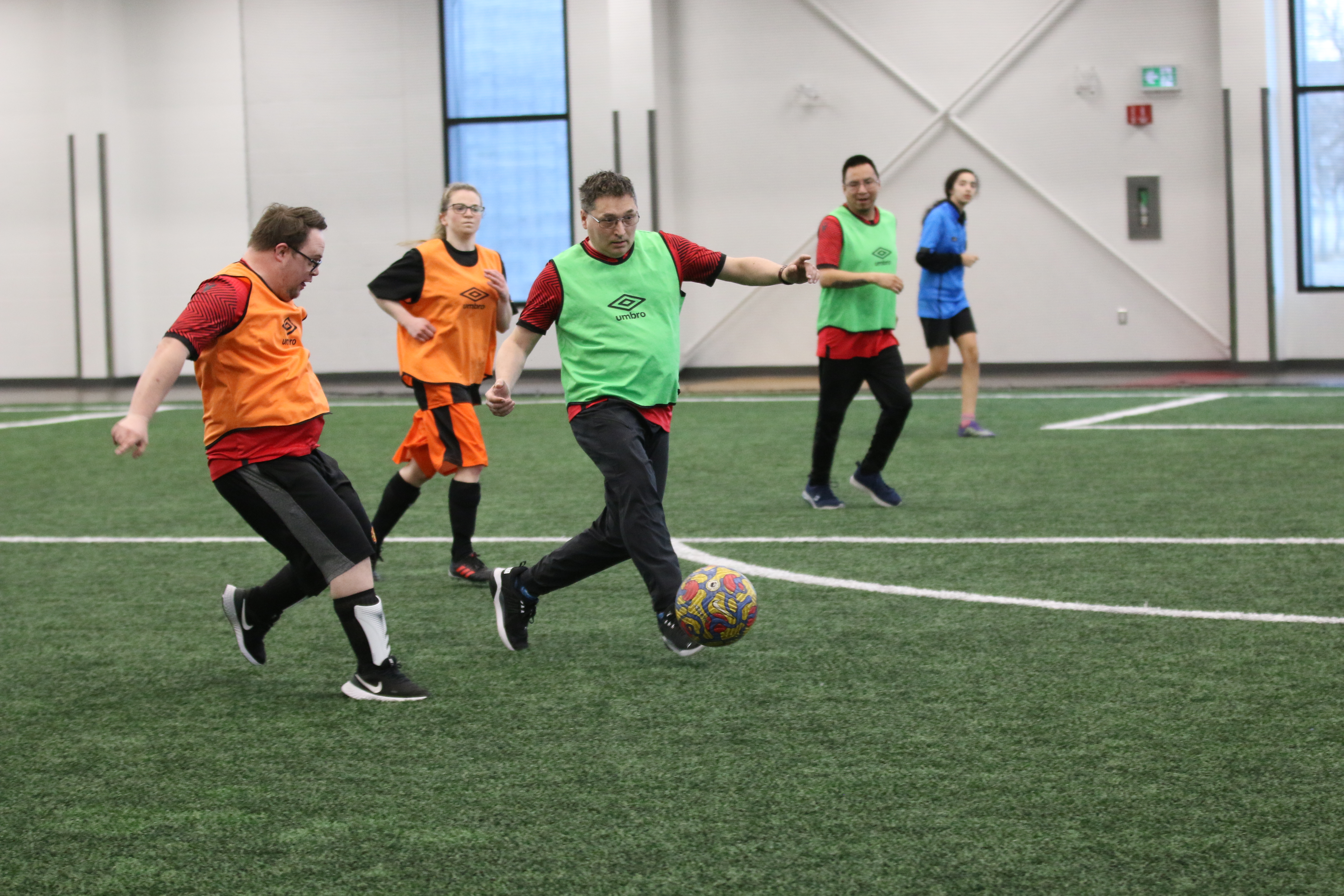 Action shot of a soccer match. Five players can be seen on the field playing soccer. One is in possession of the soccer ball. Two players are wearing orange pennies and two players are wearing light green pinnies. There is one player in a blue soccer shirt.