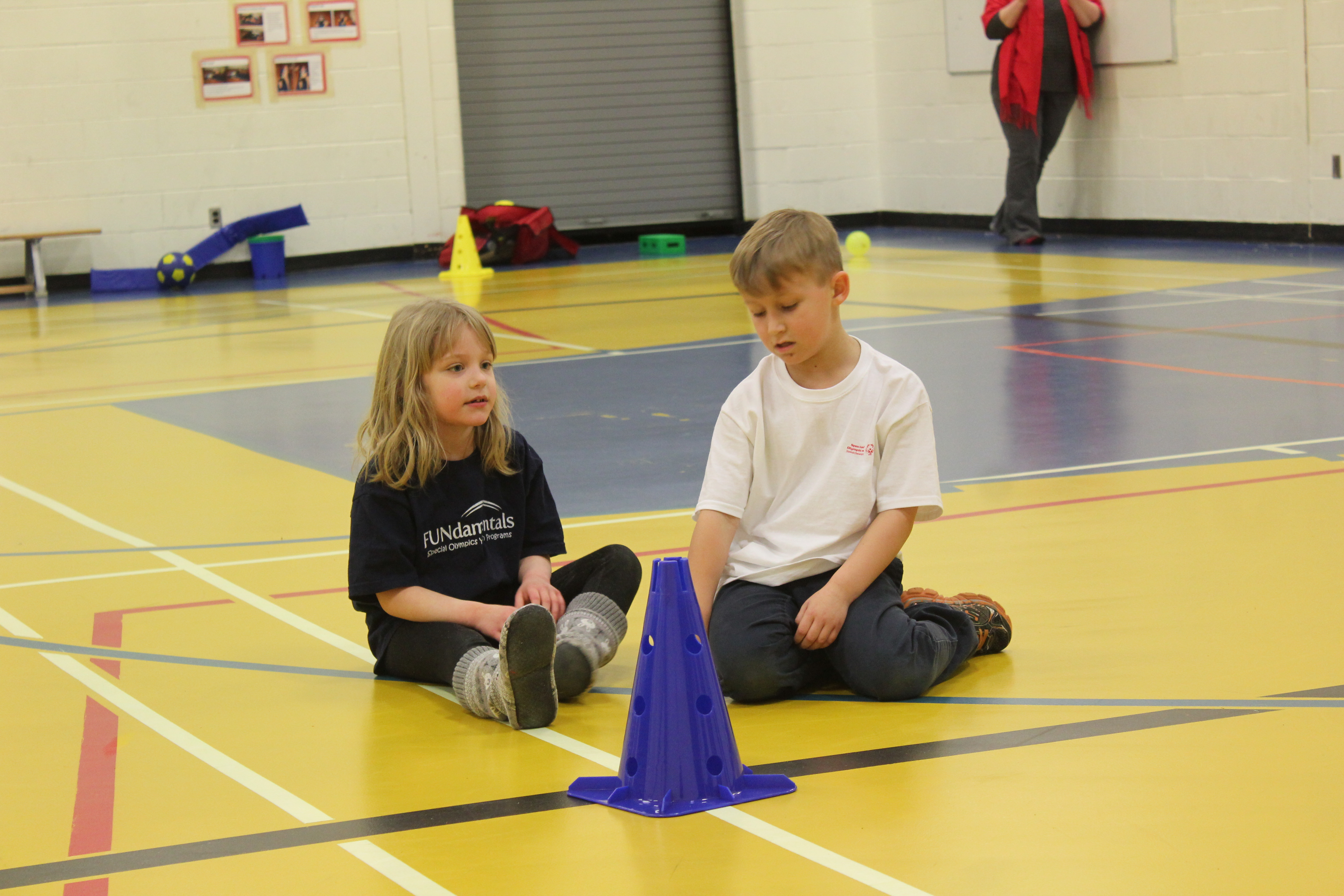 Two small children are sitting on a gym floor in front of a blue pylon. The girl on the left is wearing a black FUNdamentals t-shirt and the boy on the right is wearing a white Special Olympics Saskatchewan t-shirt.
