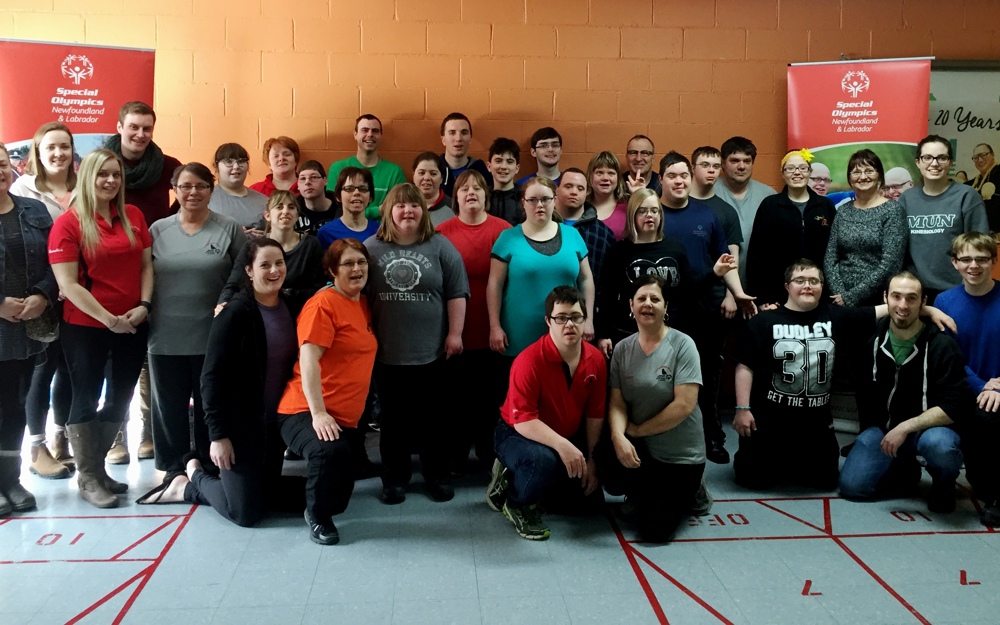 A group photo of multiple athletes, coaches, and volunteers standing against an orange brick wall. Everyone is smiling for the camera and wearing casual clothes.