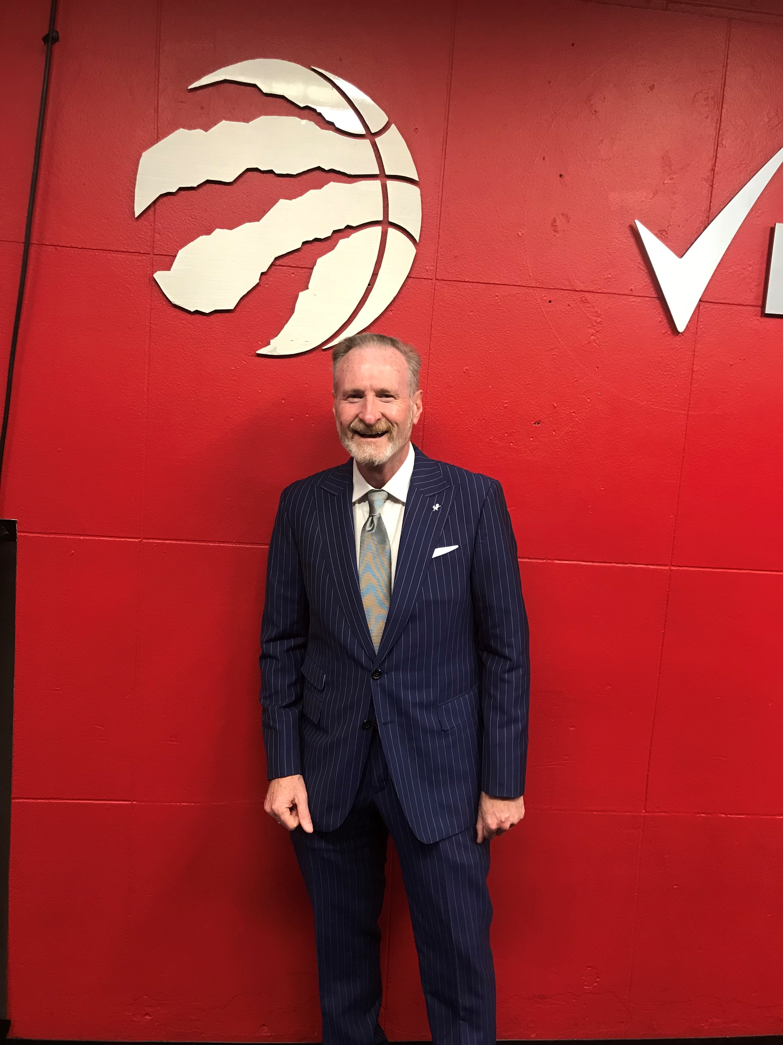 Portrait of a man in a stripped navy and white suit standing in front of a red wall with the Raports basketball logo. Portrait of a man in a stripped navy and white suit standing in front of a red wall with the Raports basketball logo.