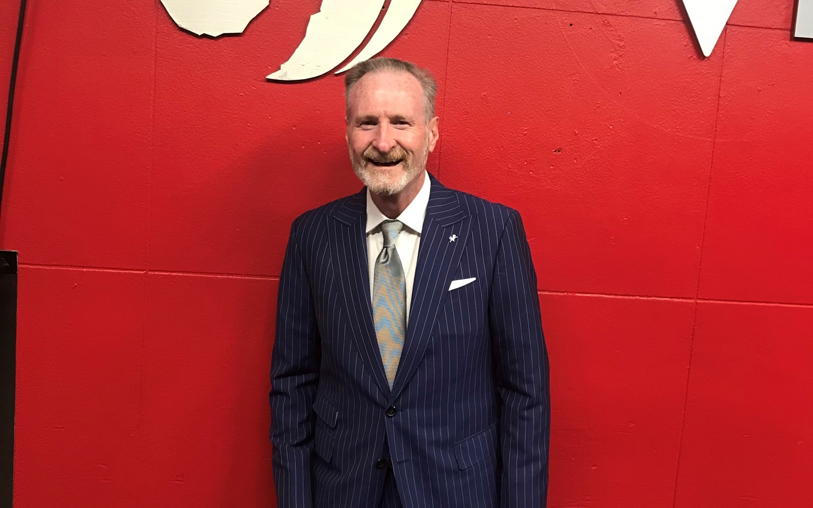 Portrait of a man in a stripped navy and white suit standing in front of a red wall with the Raports basketball logo. 