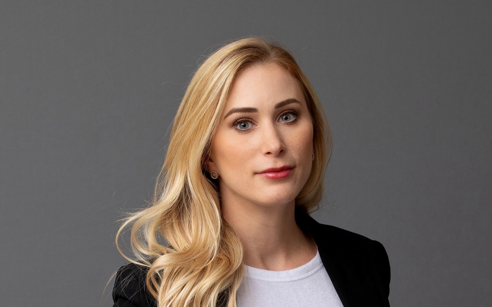 Portrait d'une femme aux longs cheveux blonds, vêtue d'un pull blanc et d'un blazer noir.