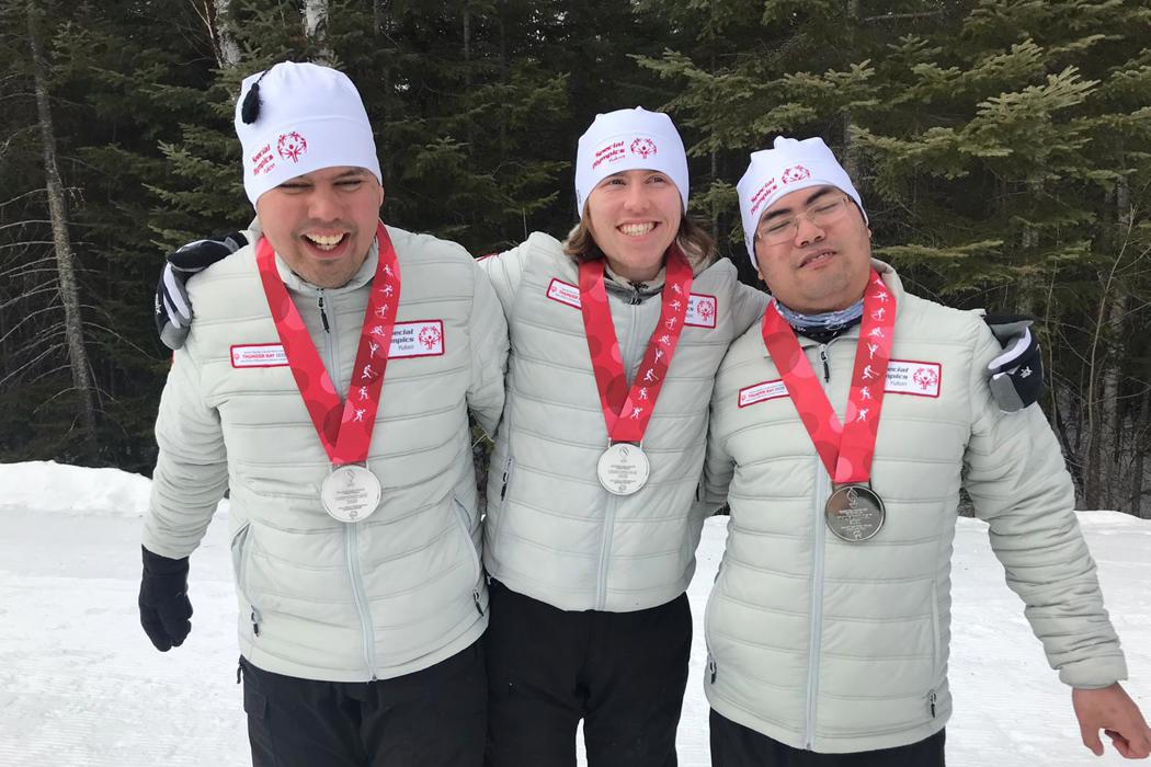 A group of three athletes hugging and smiling with silver medals around their necks. They all wear the same cream coloured jacket and white toques. There are trees in the background and snow on the floor. 