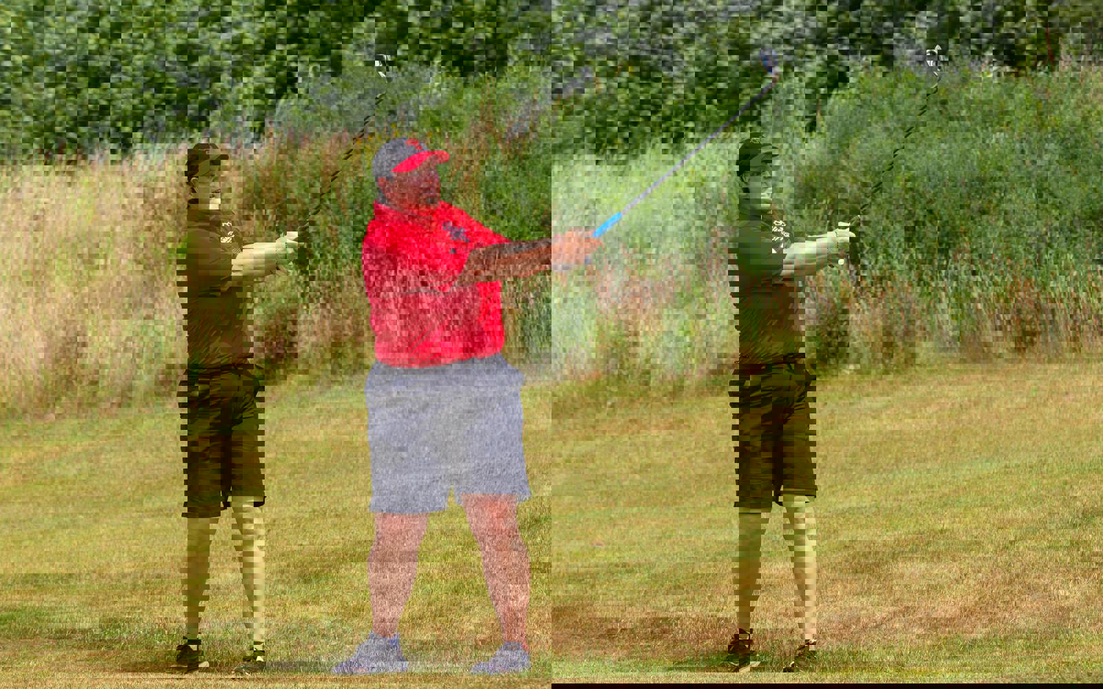 Person following through on a golf swing, dressed in a red golf shirt and hat and black shorts.