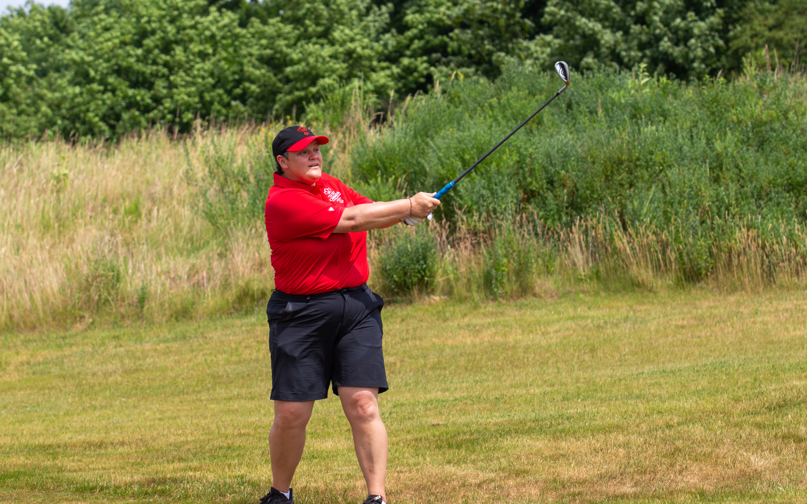 Person following through on a golf swing, dressed in a red golf shirt and hat and black shorts. 