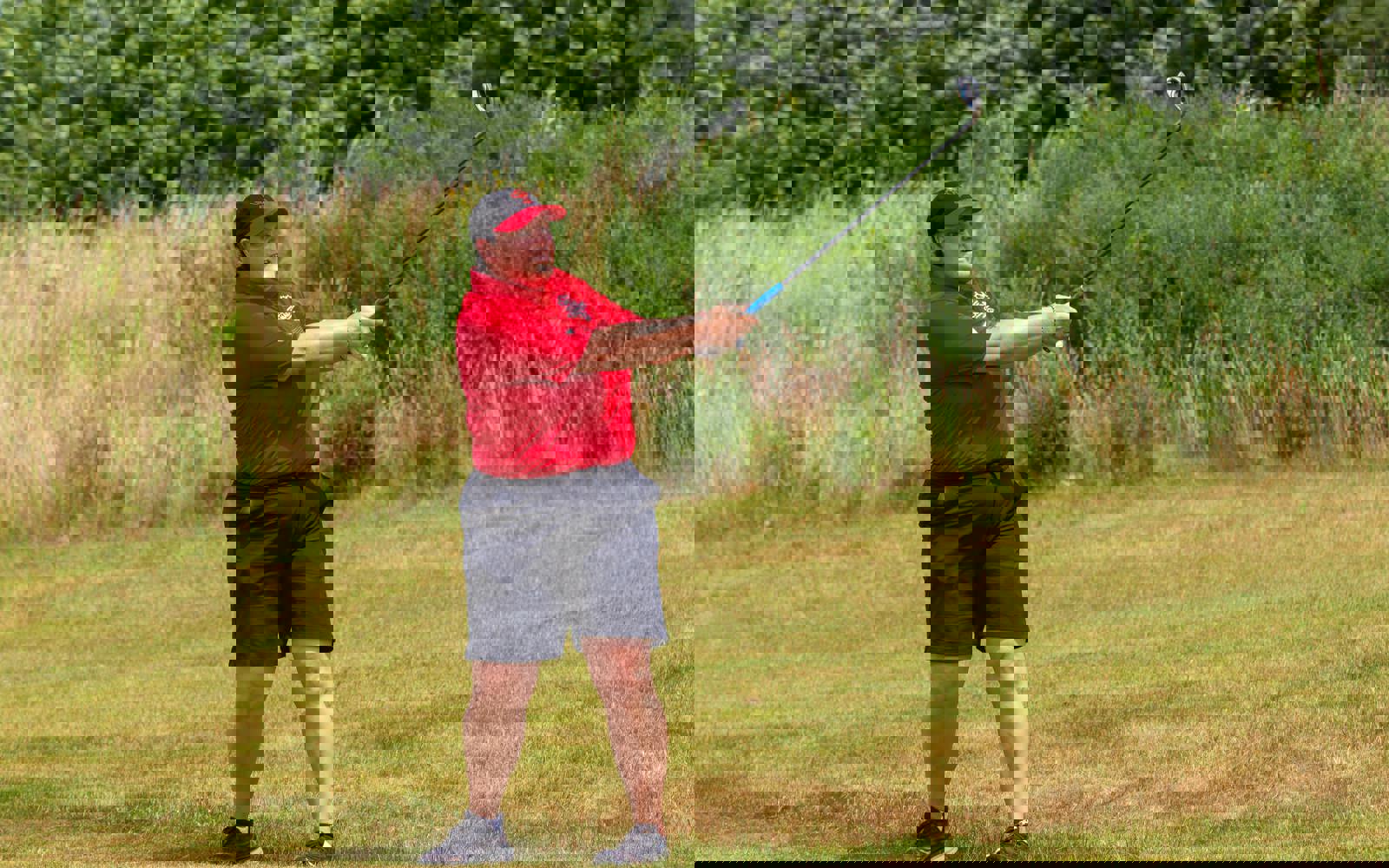 Person following through on a golf swing, dressed in a red golf shirt and hat and black shorts. 