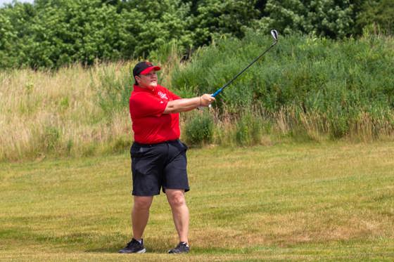 Person following through on a golf swing, dressed in a red golf shirt and hat and black shorts. 