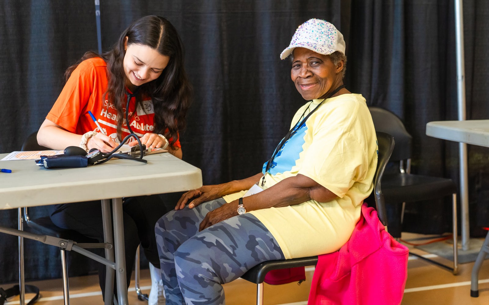 Woman in an orange tshirt writes something down while a woman in a yellow tshirt smiles at the camera.
