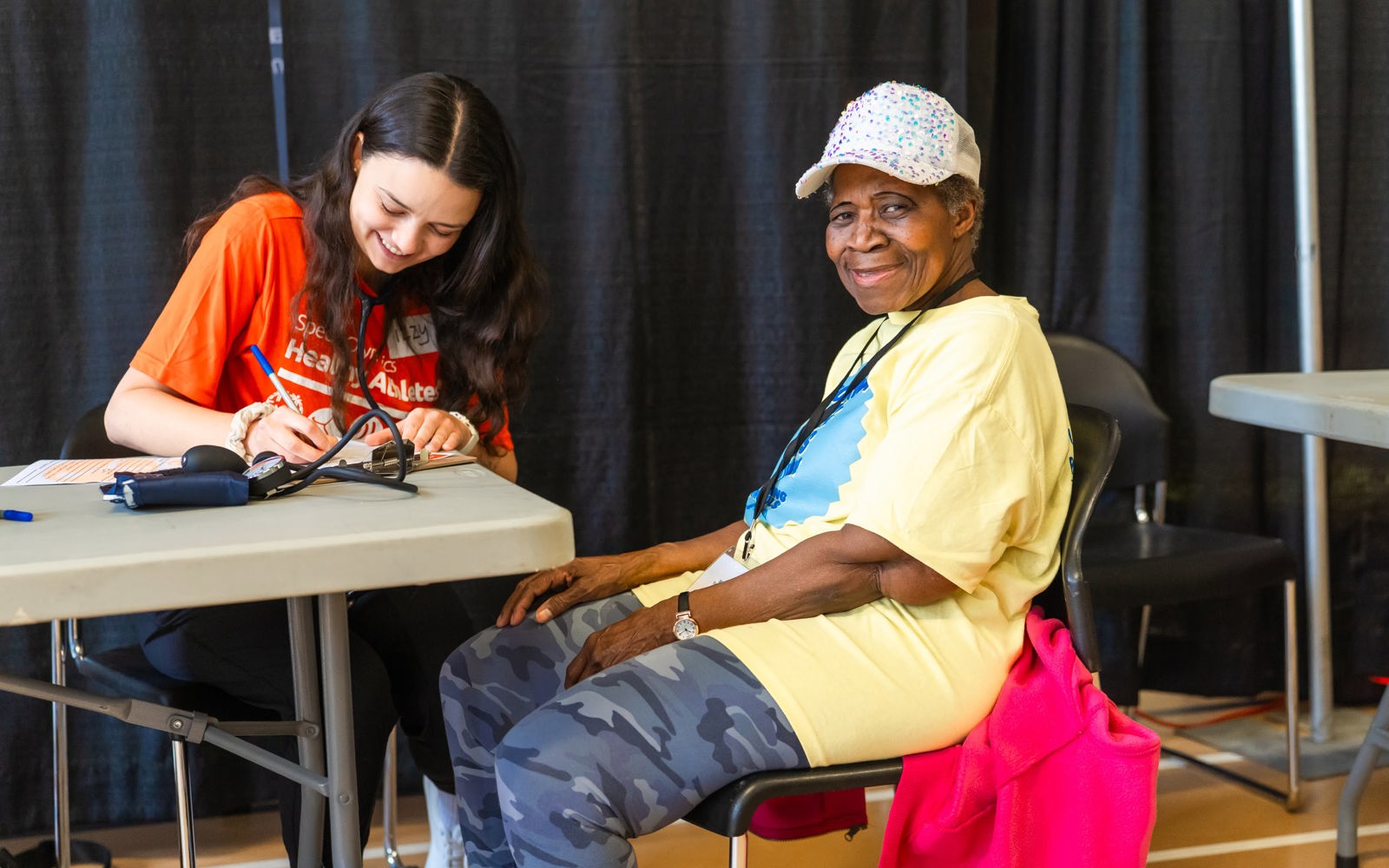 Woman in an orange tshirt writes something down while a woman in a yellow tshirt smiles at the camera. 