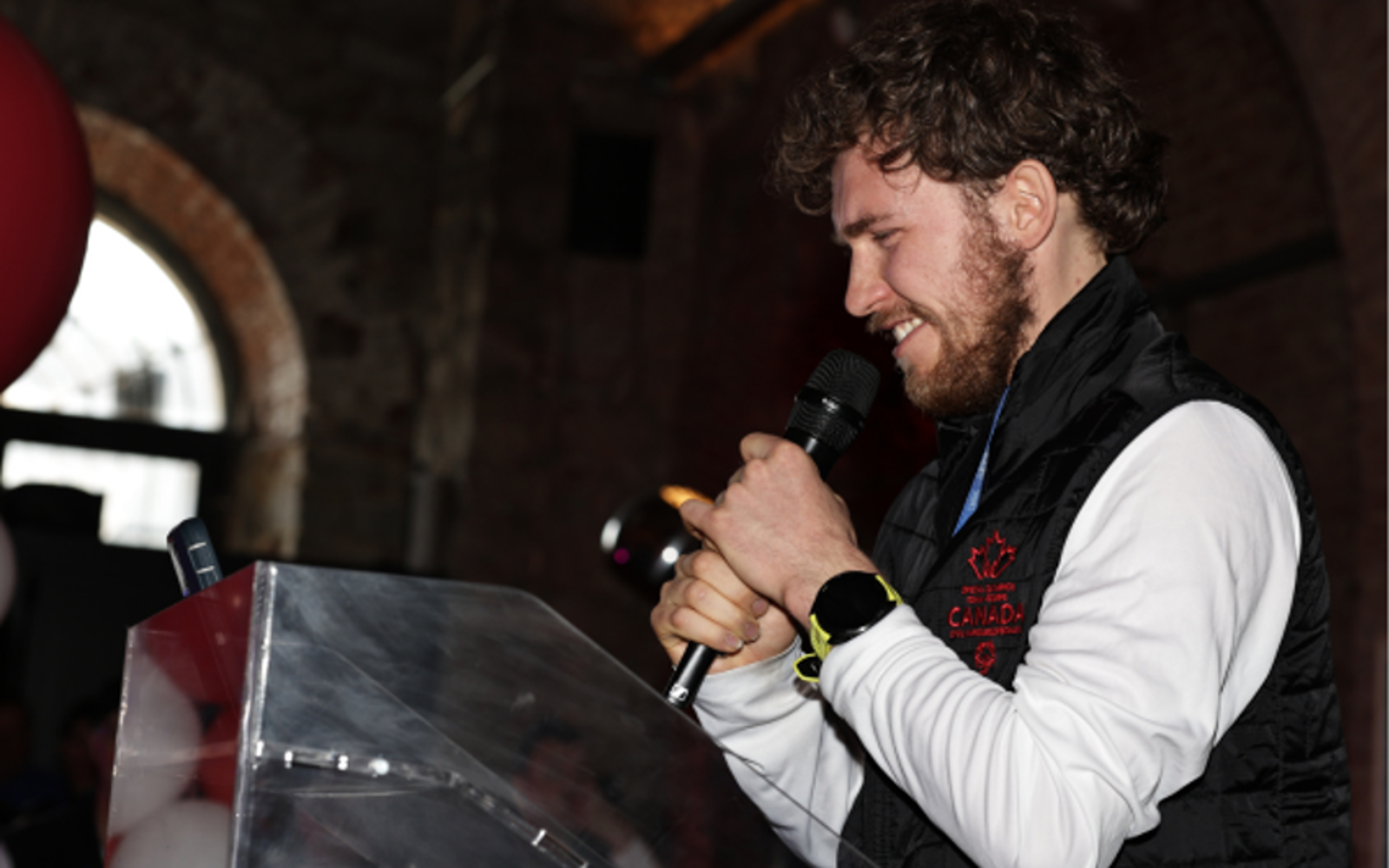 Young person in a white shirt and black vest holds a microphone close to their face. 