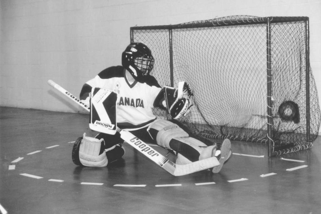 Photo en noir et blanc d’un gardien de but de l’équipe canadienne de hockey en salle, en équipement complet, portant un maillot du Canada, effectuant un arrêt contre un adversaire invisible.