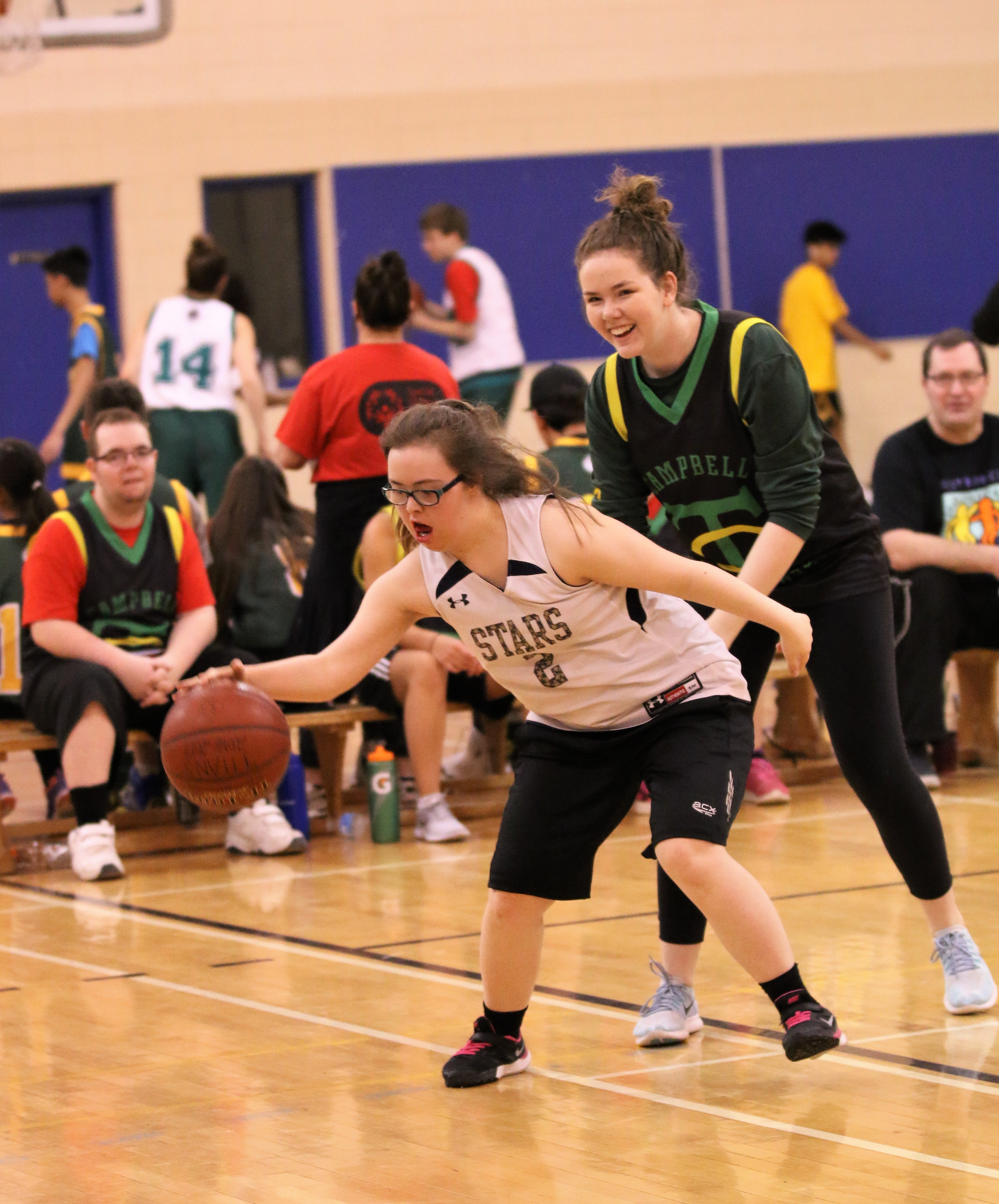 Action shot photo of two athletes playing basketball. One athlete in a white jersey has possession of the basketball. The other athlete is standing behind her and wearing a black jersey. Behind the play, you can see other athletes sitting on the bench.
