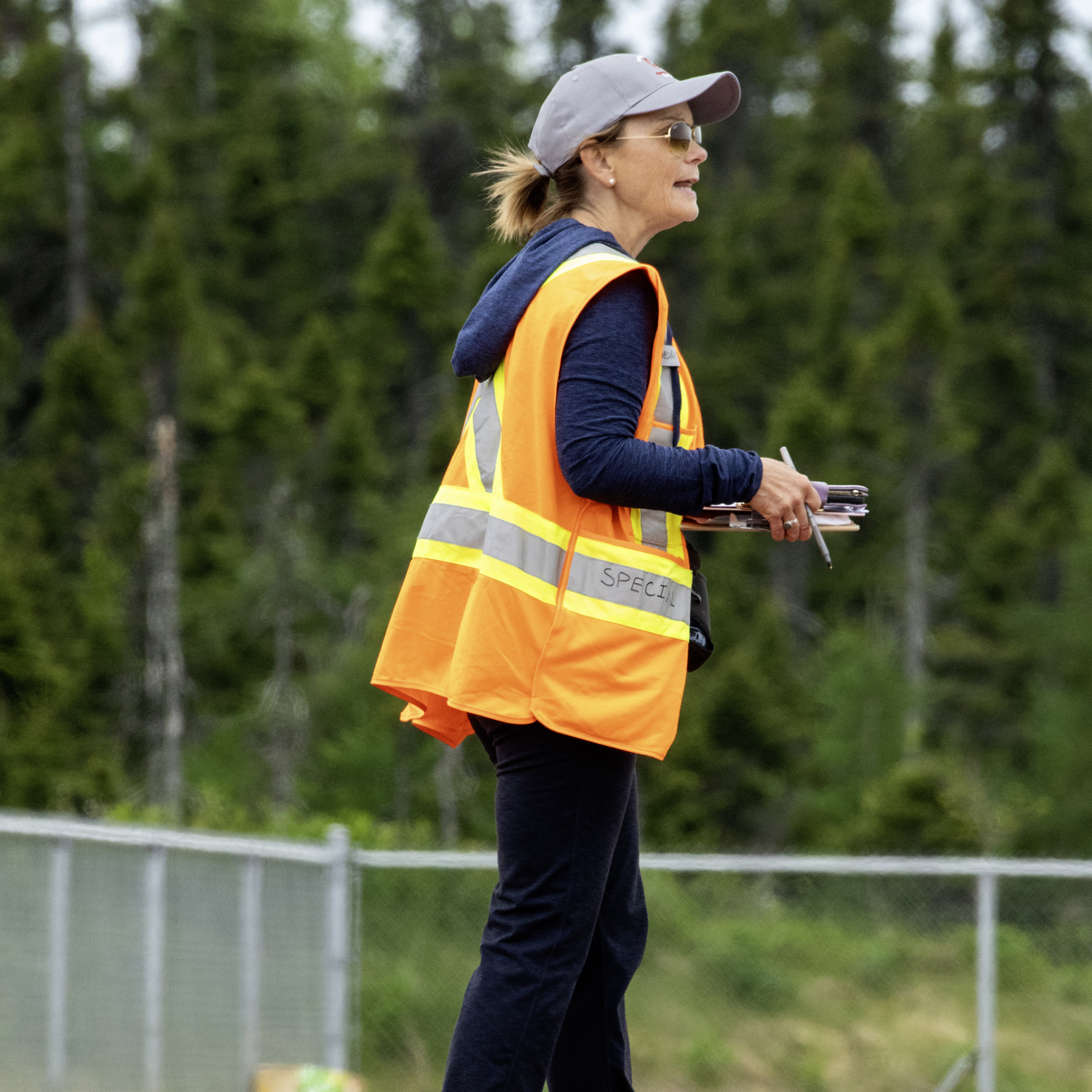 A coach, on the sideline, watching the match with her clipboard ready to take notes on what she is watching. She is wearing a high visibility vest, a baseball cap, and sunglasses.