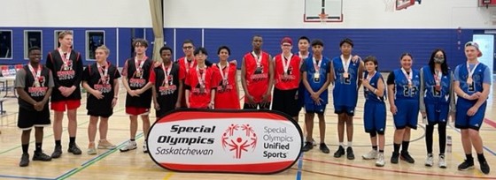Basketball athletes posing for a group photo. They are standing in a gymnasium in front of a sign that reads, "Special Olympics Saskatchewan. Special Olympics Unified Sports." Every athlete has a medal around their necks. From left to right are athletes wearing black jerseys, red jerseys, and blue jerseys.