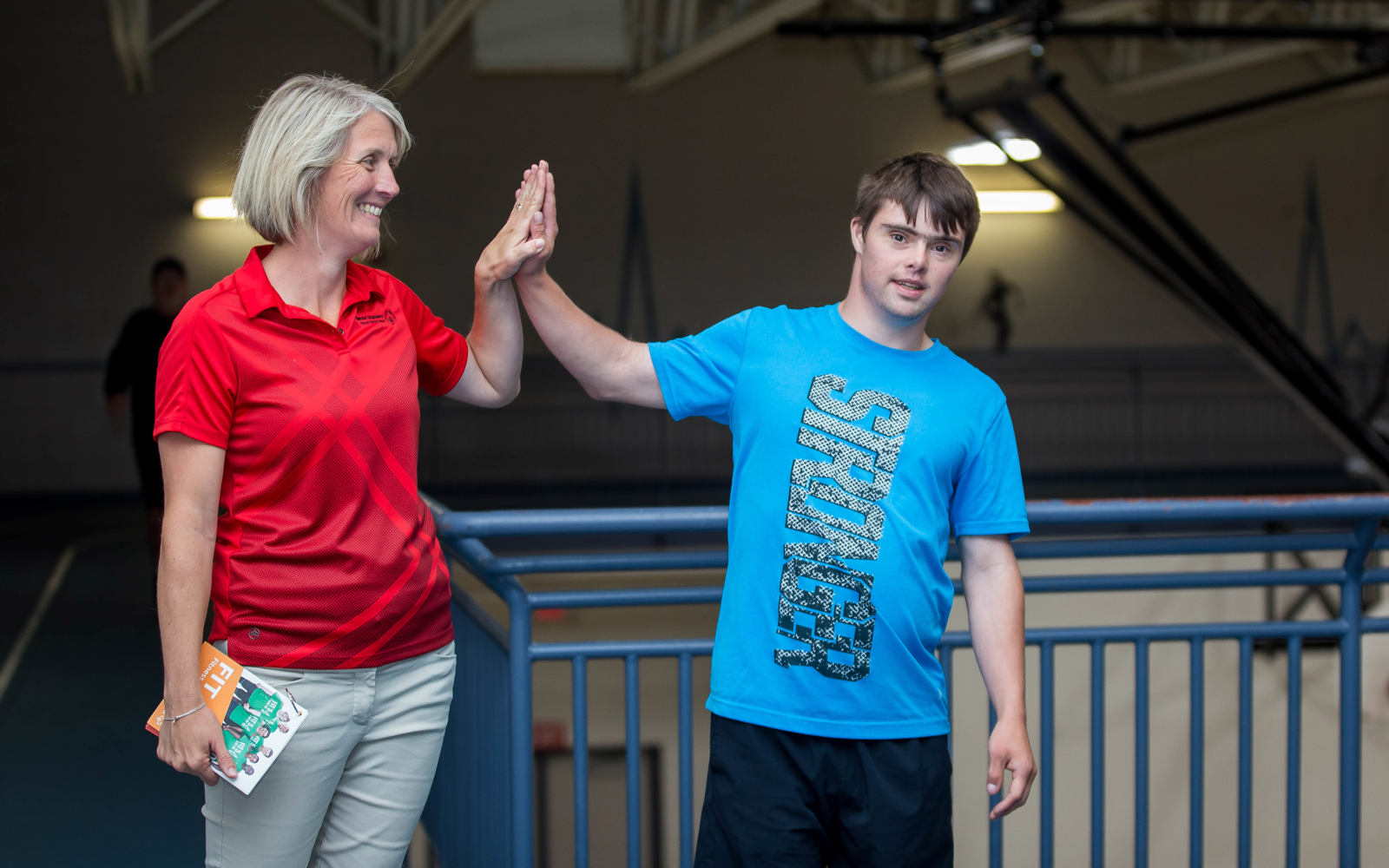 A health forum volunteer and an athlete are high-fiving one another. The volunteer is smiling and looking at the athlete while the athlete is looking at the camera. The photo takes place on the balcony of a gymnasium.