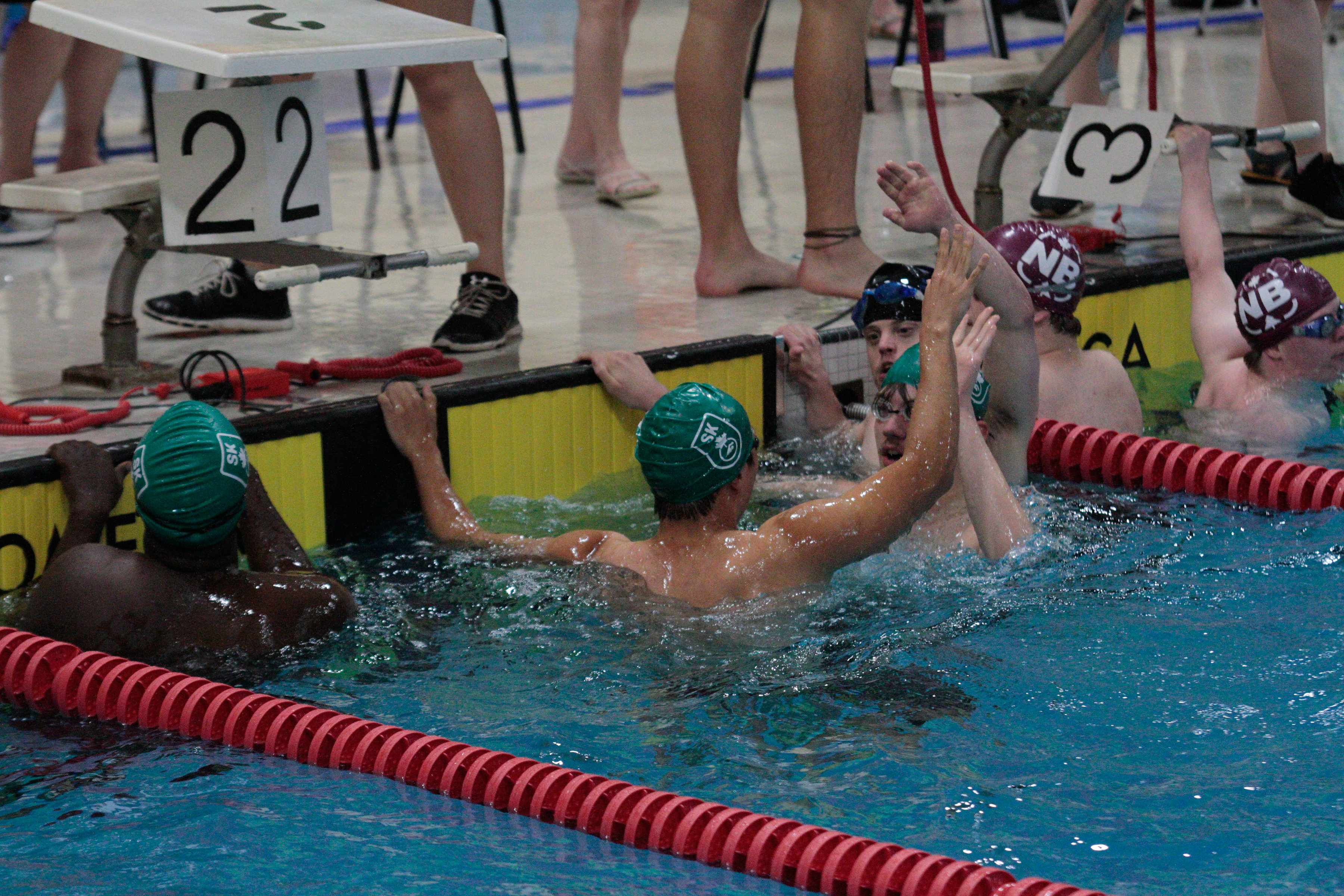 Six swimmers are holding on to the side of the pool, in the water, and giving each other high-fives. The two athletes in the front of the image have their backs turned towards the camera while you can just see the faces of the two athletes they are high-fiving. Six swimmers are holding on to the side of the pool, in the water, and giving each other high-fives. The two athletes in the front of the image have their backs turned towards the camera while you can just see the faces of the two athletes they are high-fiving.