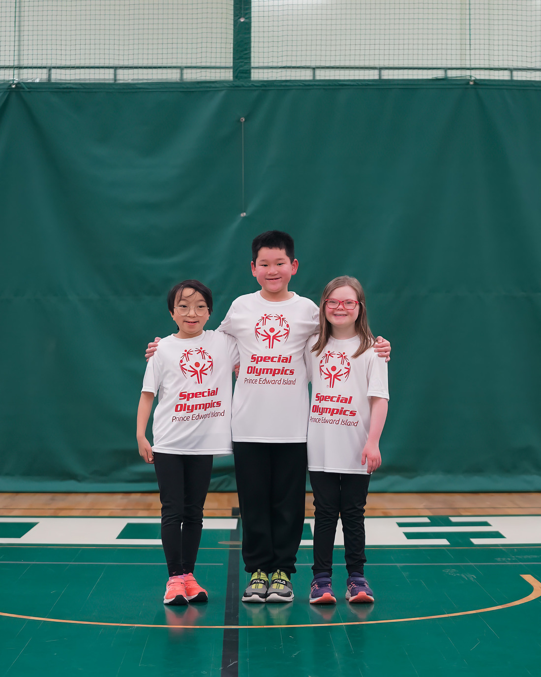 Three young kids stand in a line. All wearing Special Olympics white shirts and black pants. 