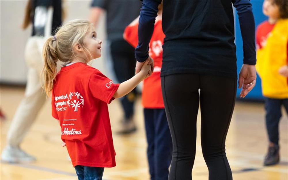Une jeune athlète regarde vers le haut en tenant la main de sa tutrice. Elle porte un chandail « Olympiques spéciaux Nouvelle-Écosse » et sourit à la personne avec qui elle se tient la main. La photo est prise de dos, et elles se trouvent dans un gymnase.