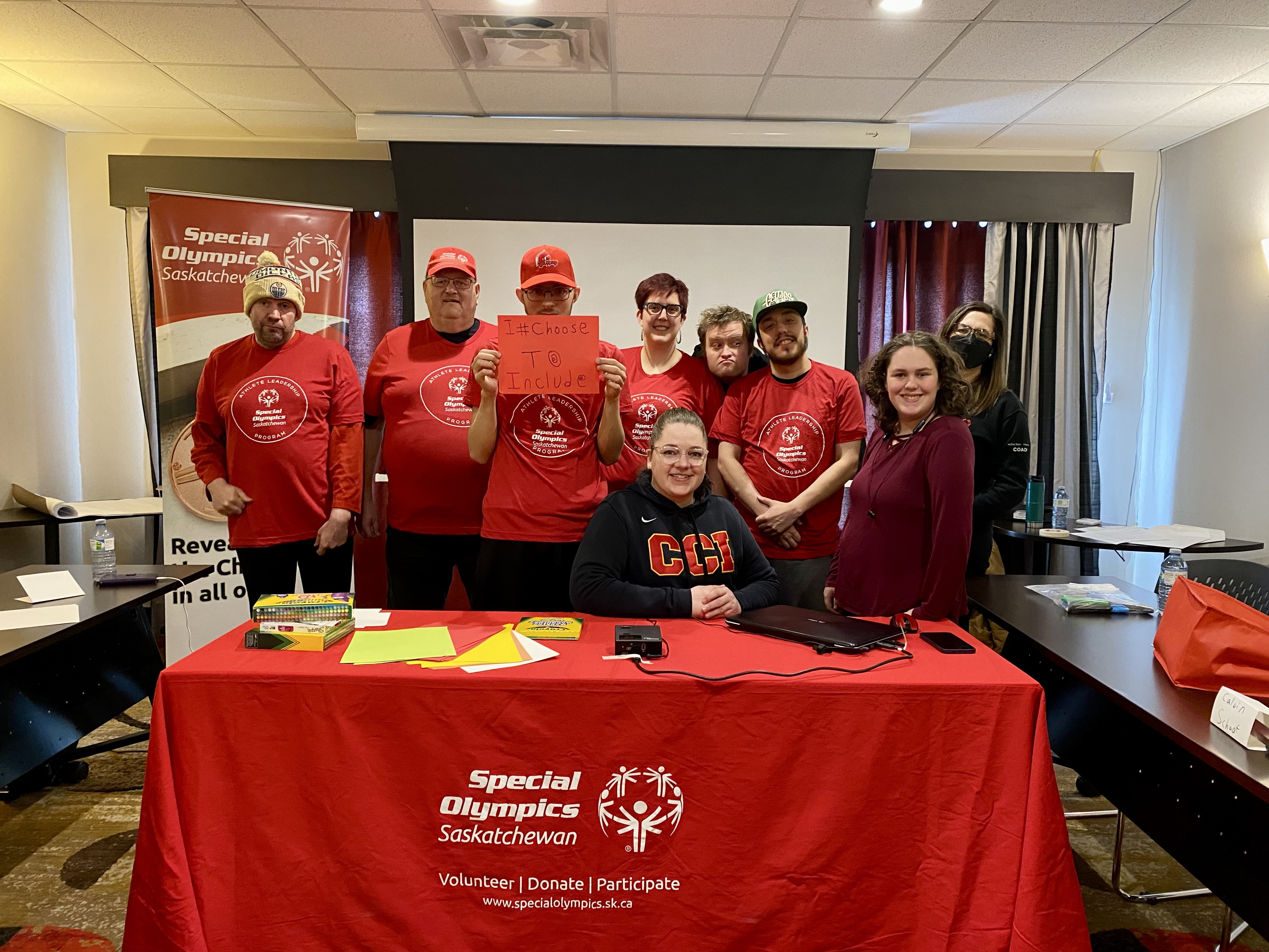 A group of athletes and volunteers are sitting and standing behind a table and smiling for the camera. Many people are wearing red Special Olympics Saskatchewan long sleeved shirts.