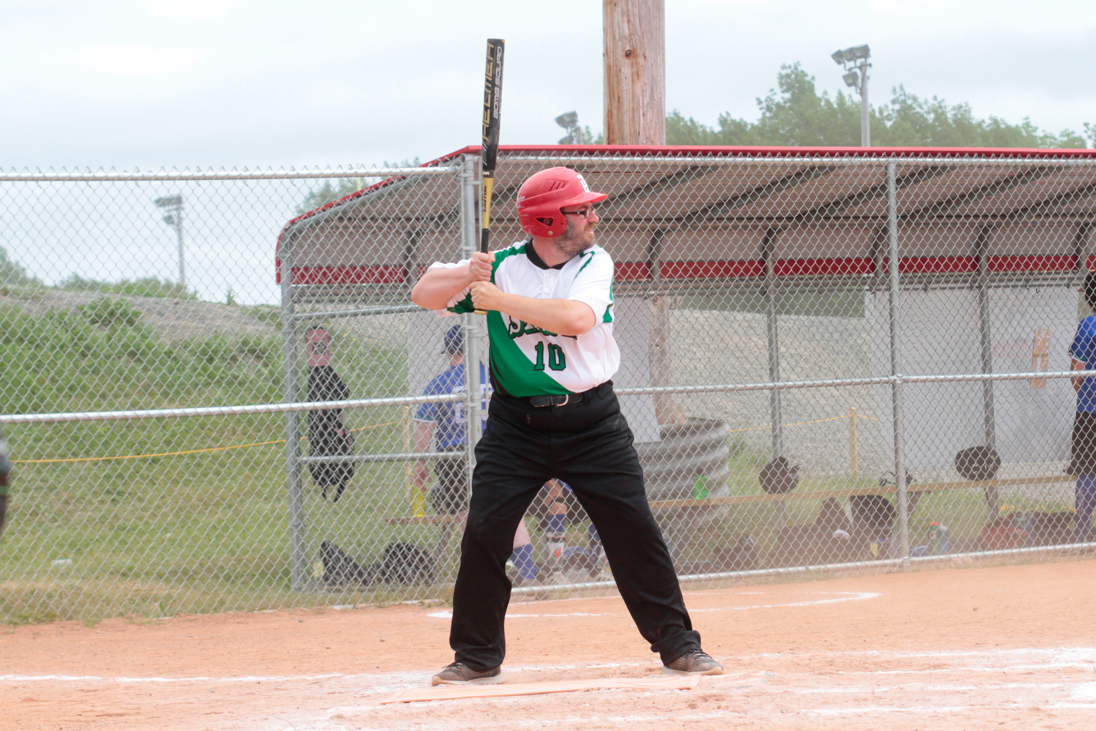 A baseball athlete at the base getting ready to hit the baseball. They are wearing a green Special Olympics Saskatchewan jersey with a red helmet. The baseball bat is swung behind their shoulder.