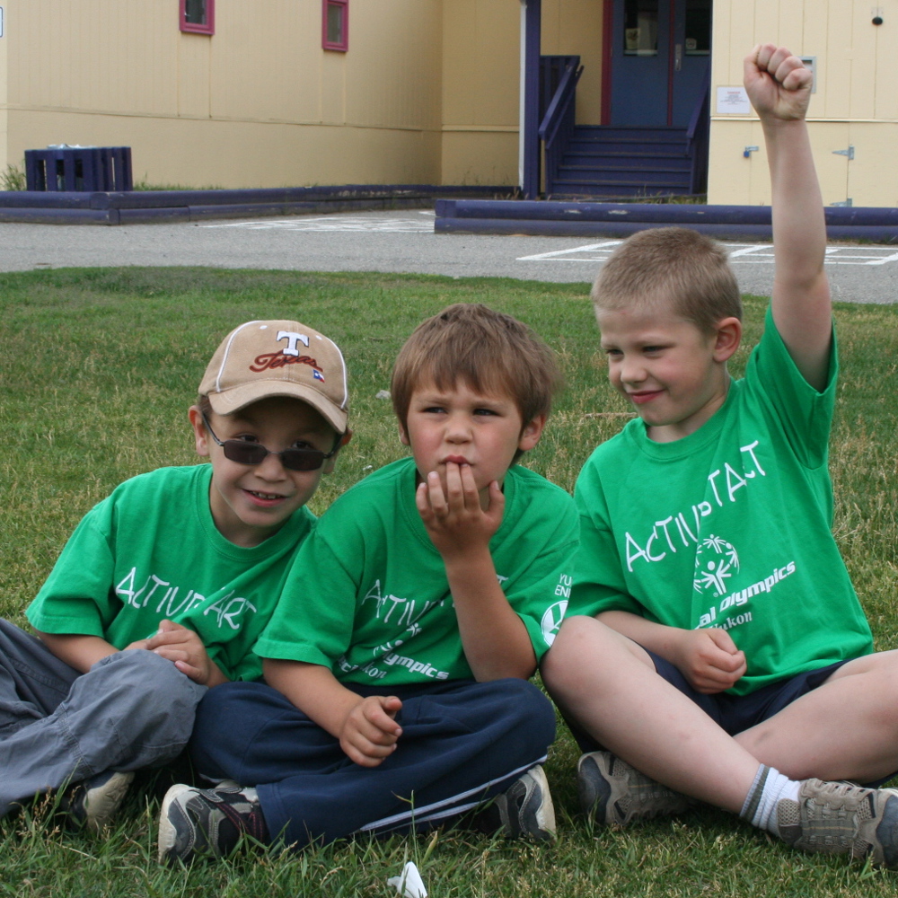 Three young athletes, wearing green Active Start t-shirts, are sitting cross-legged together in a grass field. Behind them is a school building.