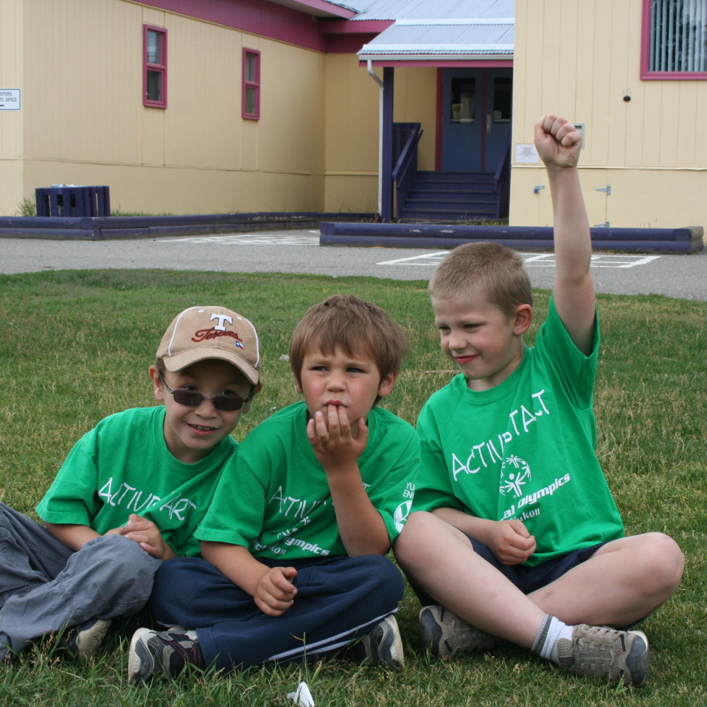 Three young athletes, wearing green Active Start t-shirts, are sitting cross-legged together in a grass field. Behind them is a school building.