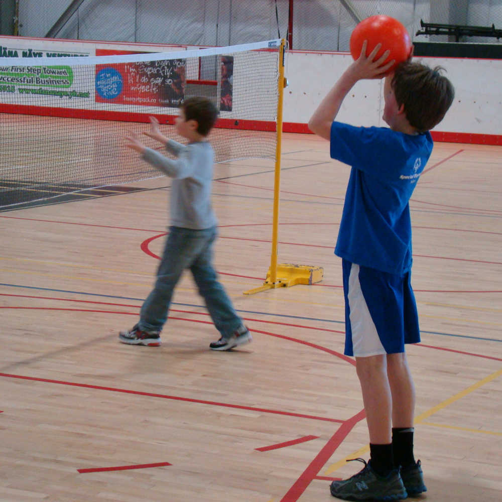 Two young athletes are playing volleyball in a gymnasium. They are standing behind a volleyball net. One boy, the boy in front is out of the camera's focus. The other boy behind him is in focus and holding up a red ball ready to throw it over the net.