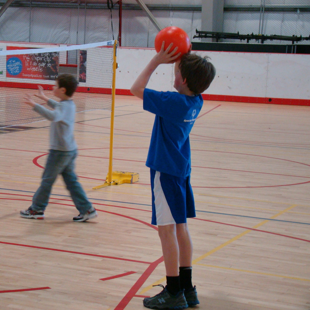 Two young athletes are playing volleyball in a gymnasium. They are standing behind a volleyball net. One boy, the boy in front is out of the camera's focus. The other boy behind him is in focus and holding up a red ball ready to throw it over the net.
