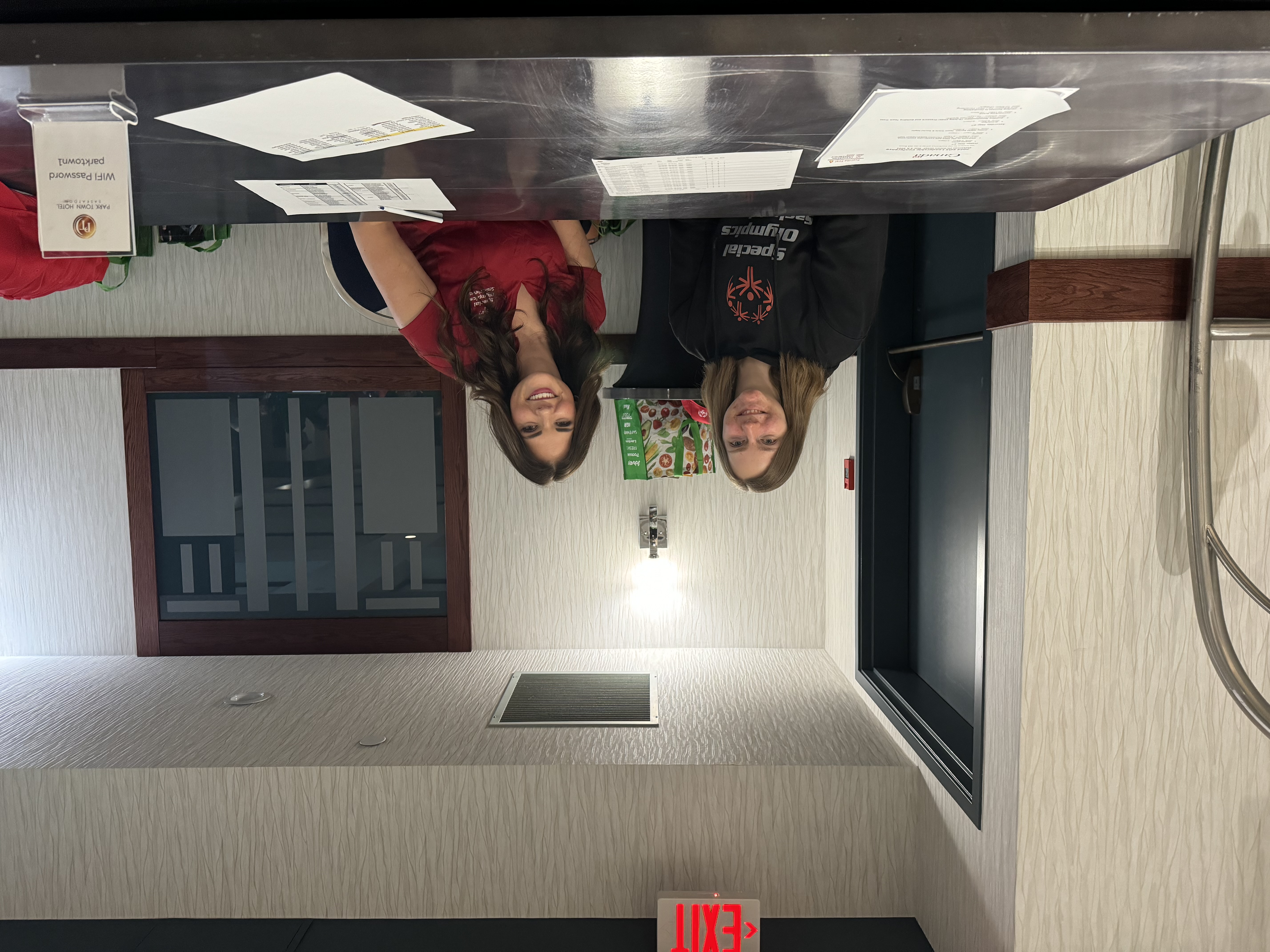 Two volunteers are sitting behind a table and smiling for the camera. In front of them are stacks of paper.