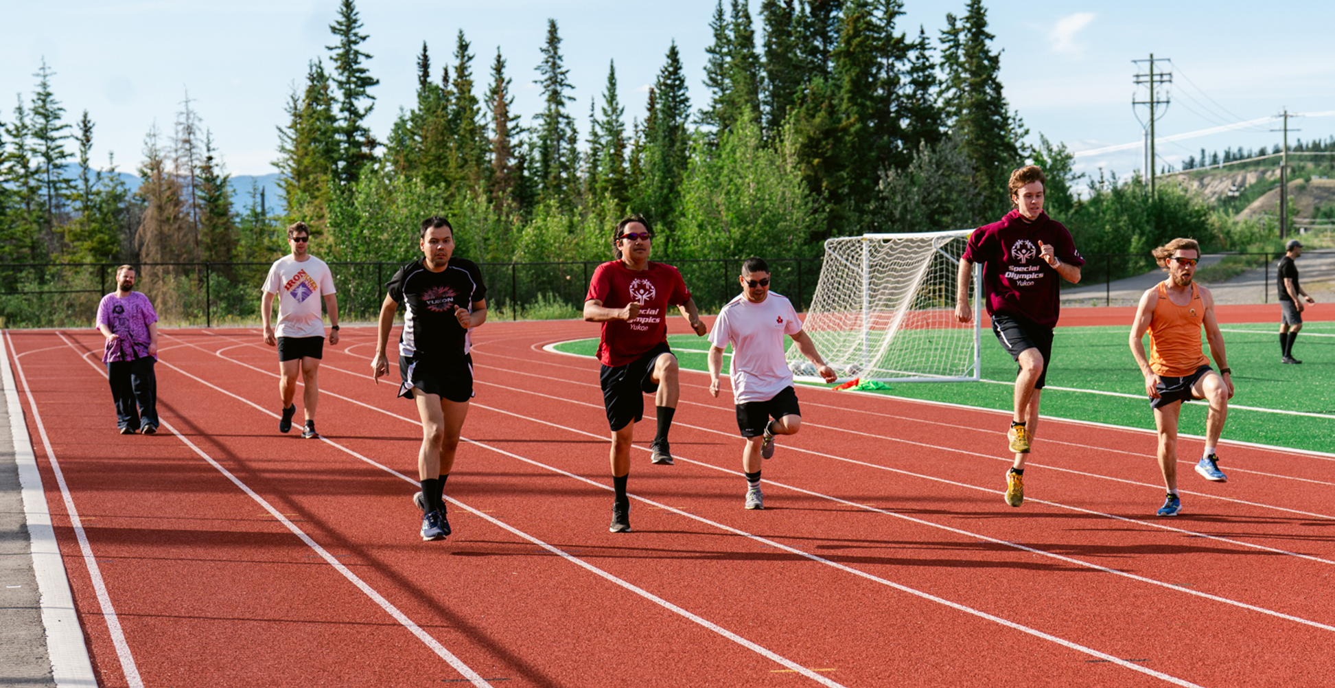 Seven athletes running along a track. It's a sunny day and the athletes are all wearing athleisure.